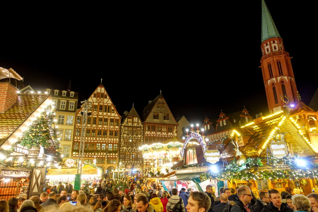 Ein gesch├Ąftiges Weihnachtsmarkt in Nürnberg, Deutschland mit Menschen um dekorierte St├Ąnde, festliche Lichter, Geb├Ąude mit Fenstern, ein Uhrturm und einen dunklen Himmel.