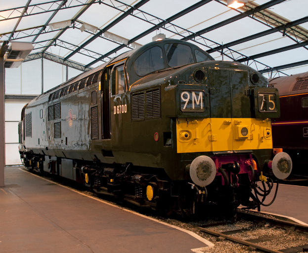 Ein Zug fährt auf den Schienen neben einem Bahnsteig mit einem an der Decke befestigten Mast, der Metallstangen mit Lampen trägt.