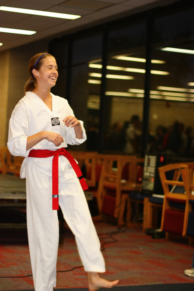 Ein Mädchen in einem Karate-Outfit mit einem roten Gürtel lacht, etwas anschauend, mit Stühlen, einer Musikanlage und ein paar Menschen hinter ihr, vor einer Wand mit Lichtern.