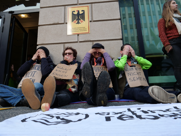 Eine Gruppe von Menschen sitzt vor einem Gebäude auf dem Boden und hält Schilder, mit einem Banner auf der Straße und einer Frau rechts daneben, offenbar bei einer Protestaktion gegen eine Regierungsentscheidung.