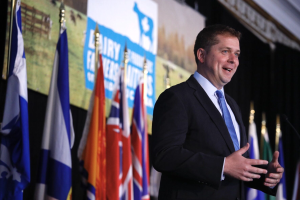 Ein Mann im Anzug spricht an einem Podium mit Mikrofonen, umgeben von Fahnen und einem Bildschirm, wahrscheinlich zu einer Regierungszahlung von 1,5 Milliarden Dollar für die Milchindustrie.