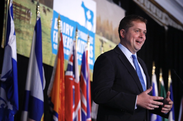 Ein Mann im Anzug spricht an einem Podium mit Mikrofonen, umgeben von Fahnen und einem Bildschirm, wahrscheinlich zu einer Regierungszahlung von 1,5 Milliarden Dollar für die Milchindustrie.