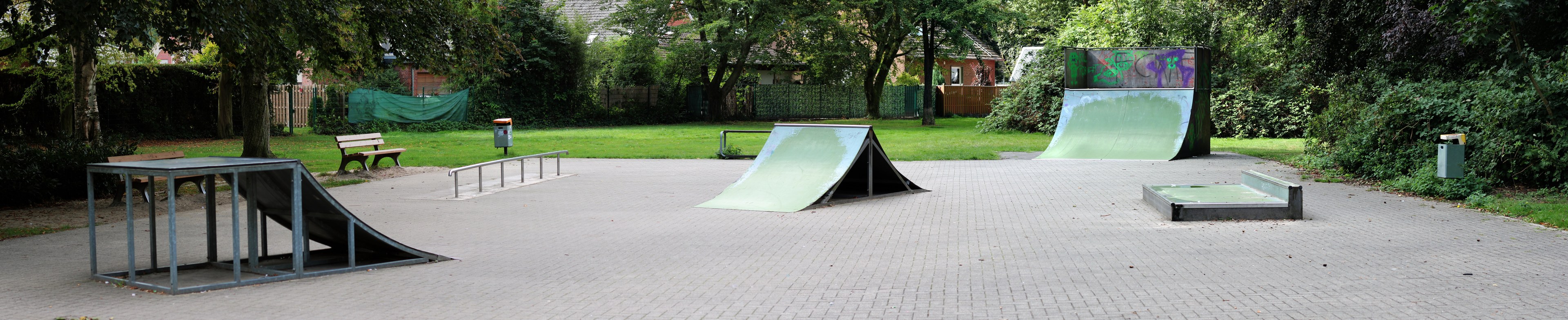 Ein Park mit einer Skateboard-Rampe in der Mitte, umgeben von Bänken, Geländern, Gras, Pflanzen, Bäumen und Gebäuden im Hintergrund.