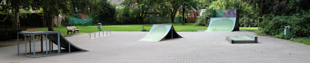 Ein Park mit einer Skateboard-Rampe in der Mitte, umgeben von Bänken, Geländern, Gras, Pflanzen, Bäumen und Gebäuden im Hintergrund.