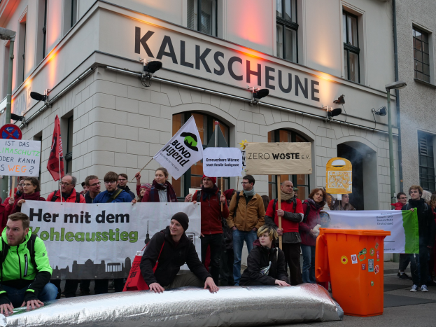 Eine Gruppe von Menschen mit Protestschildern und Plakaten vor einem Gebäude, mit zwei Personen im Vordergrund sitzend und einem Müllcontainer rechts daneben, vor einem Hintergrund von Gebäuden mit Fenstern und Schildern in Deutschland.