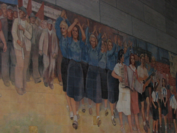 Eine Gruppe von Menschen versammelt sich, mit blau gekleideten Mädchen in der Mitte, die klatschen, Musikern auf der rechten Seite, spielenden Kindern in der rechten Ecke und Individuals mit roten Fahnen in der linken Ecke.