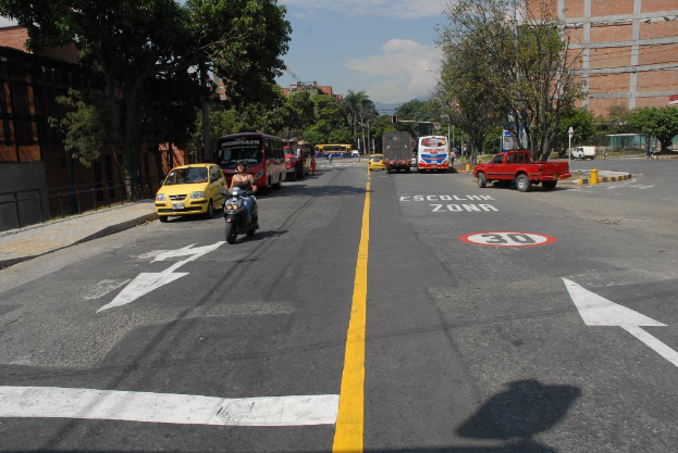 Eine Straßenszene mit Gebäuden, Bäumen, fahrenden Fahrzeugen, Schildern, Verkehrsampeln auf Pfählen, einem blauen bewölkten Himmel und Warnschildern auf der Straße.