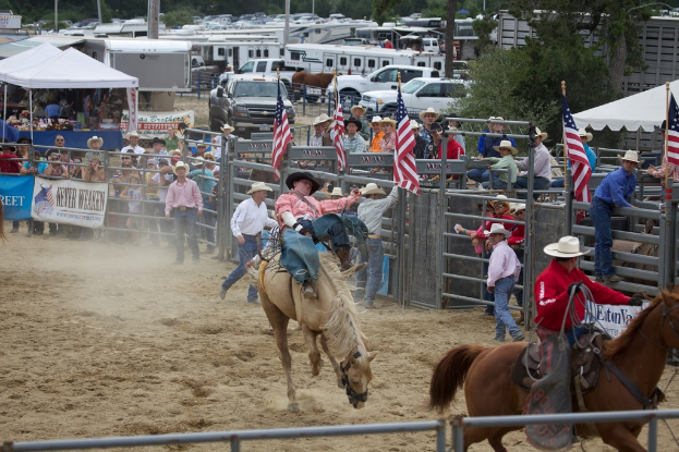 Zwei Reiter auf Pferden bei einem Wettbewerb namens Charreada, umgeben von Gruppen von Menschen, Banner und Fahnen, Zeltplanen, geparkten Fahrzeugen und Bäumen im Hintergrund.
