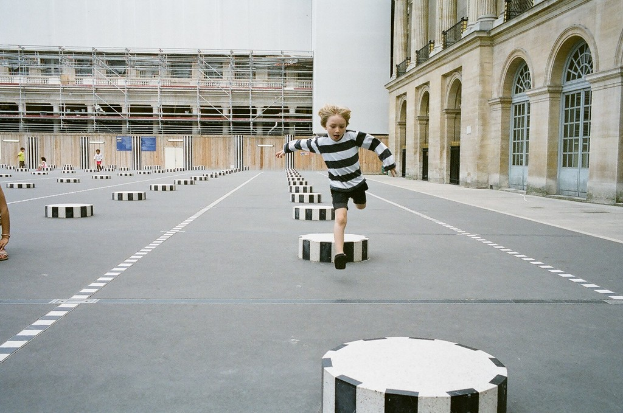 Ein Spielplatz mit vier Kindern, eines läuft auf einer schwarz-weißen Betonstufe, umgeben von einem Schulgebäude.