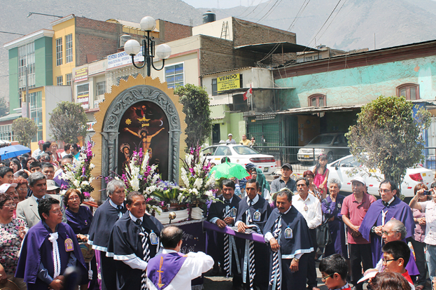 Menschen auf einer Straße außerhalb der Stadt, mit einem Kreuz und Blumensträußen in der Mitte, fahrende Autos im Hintergrund und Gebäude, Bäume und Straßenlaternen weiter hinten.
