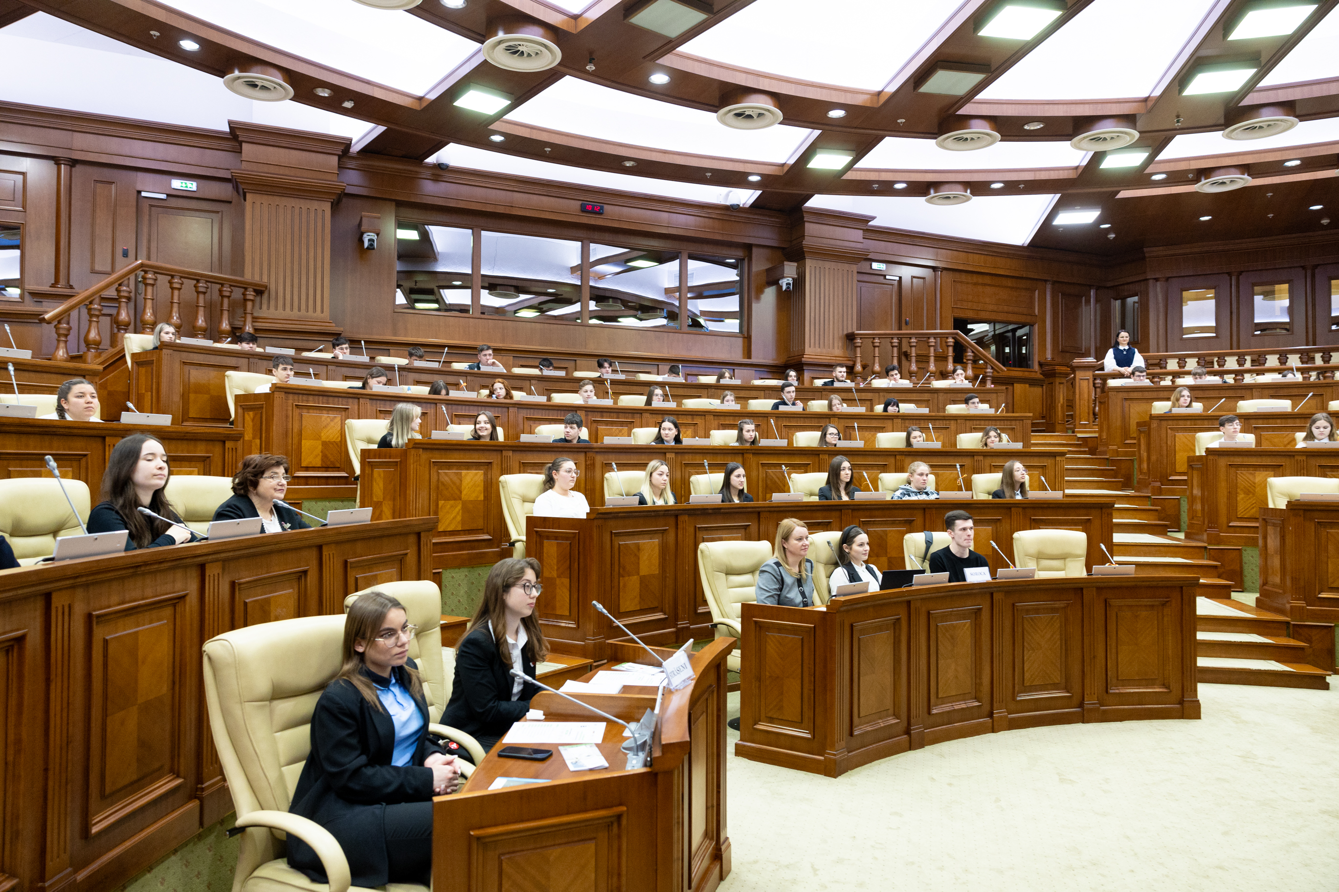 Eine große Gruppe von Menschen sitzt an Tischen in einem Raum, ausgestattet mit Mikrofonen und Papieren, an einer Sitzung des griechischen Parlaments teilnehmend, mit Treppen und Geländern im Hintergrund und Deckenbeleuchtung.