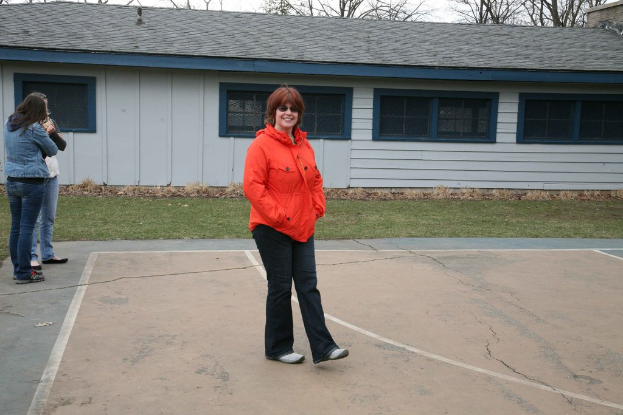 Zwei Personen auf einem Platz mit einer Frau in einem orangen Hemd, die in der Mitte läuft, Gras neben dem Platz, ein Gebäude mit einem Dach und Fenstern, Bäume und Himmel im Hintergrund.
