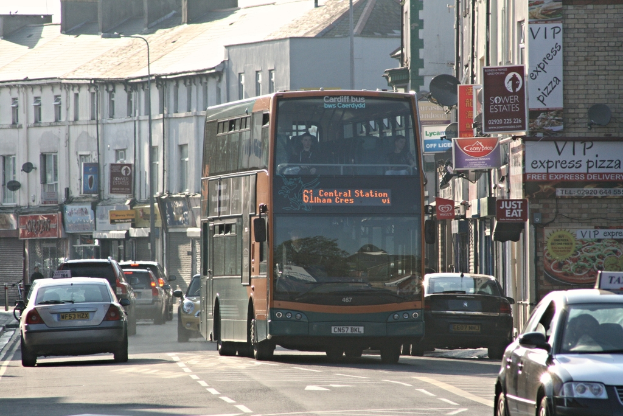 Eine Straße mit Autos und einem Bus, Gebäude mit Wänden, Fenstern, Tellern und Dächern, Plakate und Banner an den Wänden und ein Pfosten mit einer Straßeleuchte.