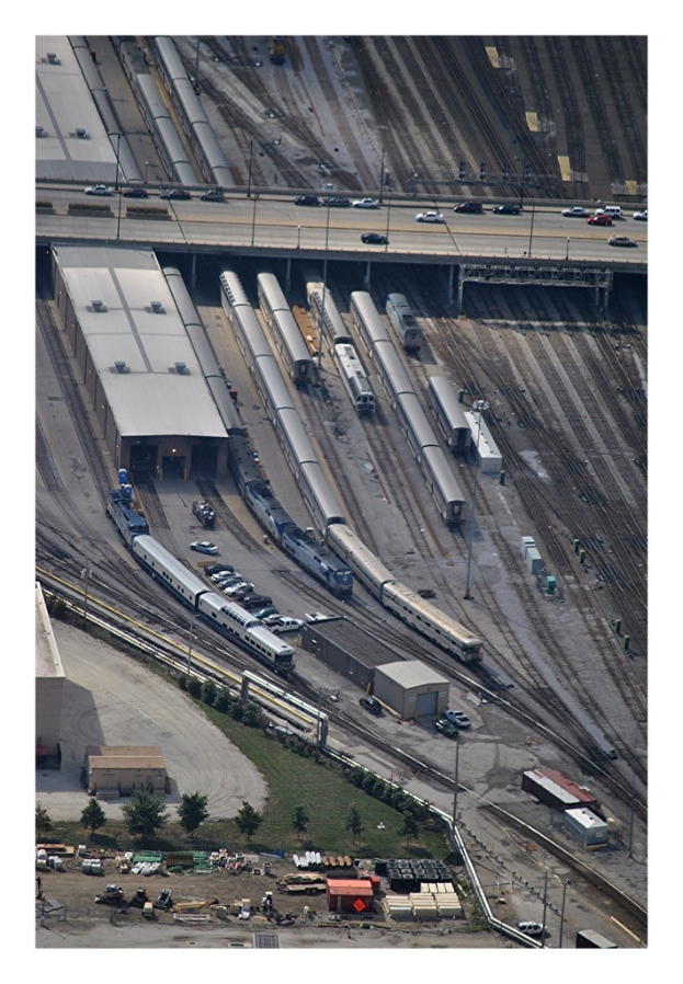 Eine Brücke mit wenigen Fahrzeugen darauf, darunter Eisenbahnschienen mit ein paar Zügen und eine Landschaft mit Gebäuden, Bäumen, Pfählen und diversen Gegenständen und Fahrzeugen am Boden.