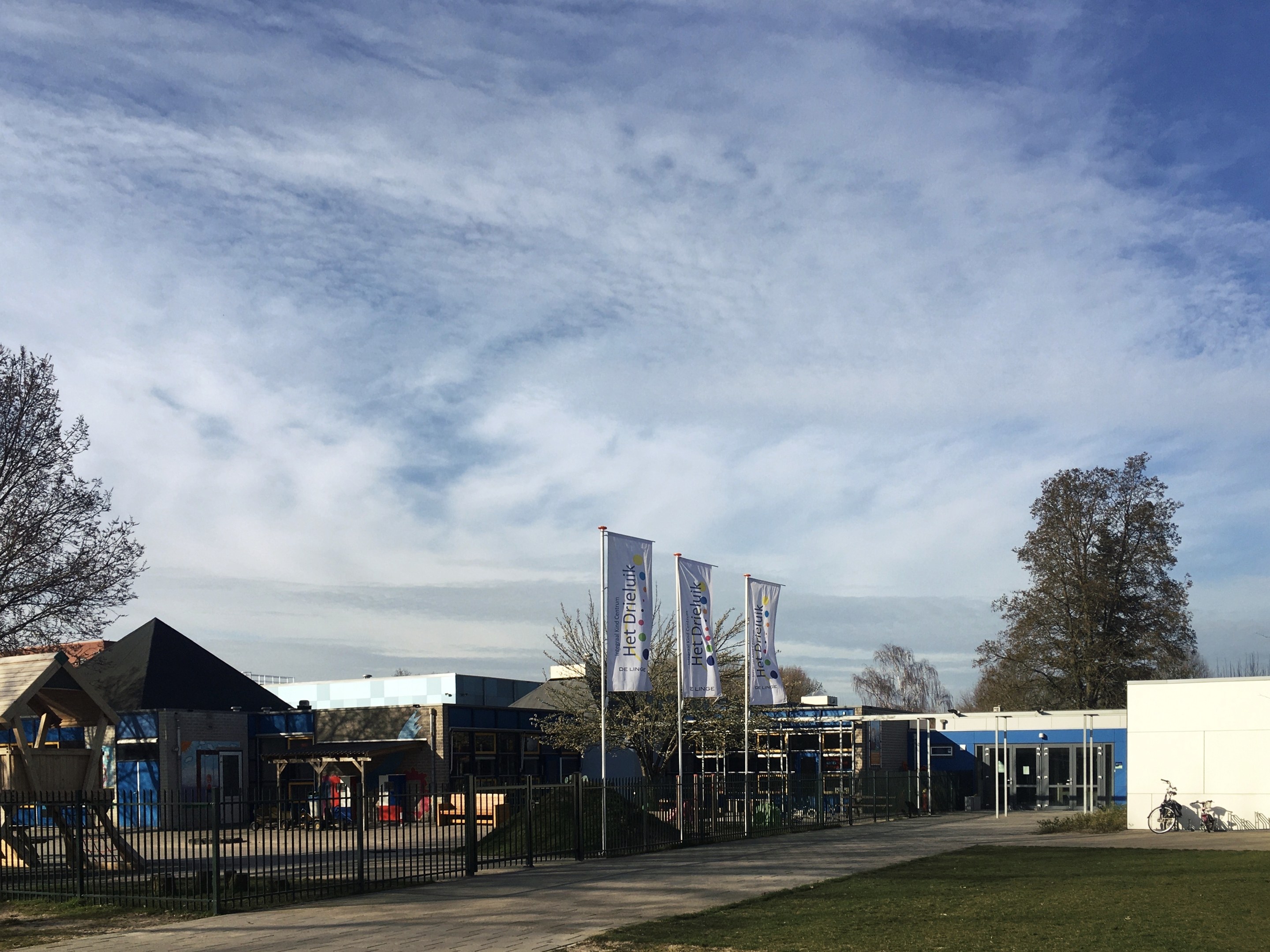 Großes Gebäude mit Metallzaun, Rasenfläche, Weg, Bäumen, Bannern, Fahrrad, nahen Häusern und bewölktem Himmel, als neuer Spielbereich an der Schule identifiziert.