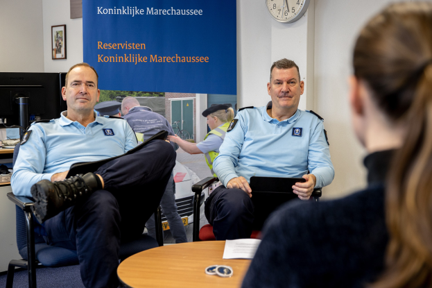 Zwei Polizeibeamte sitzen an einem Tisch mit einer Frau, umgeben von Papieren und Gegenständen, mit einem wandmontierten Banner, einer Uhr und Fotorahmen im Hintergrund und einem zweiten Tisch mit einem Monitor und Zubehör links daneben.
