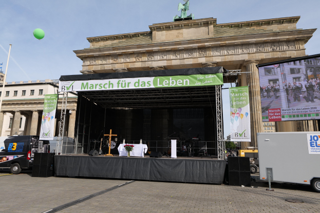 Eine Bühne vor dem Brandenburg Tor in Berlin, Deutschland, mit einem Tisch mit einer Vase, einem Kreuzsymbol, Bannern, Lautsprechern und anderen Gegenständen, sowie Fahrzeugen auf der Straße und Gebäuden im Hintergrund.