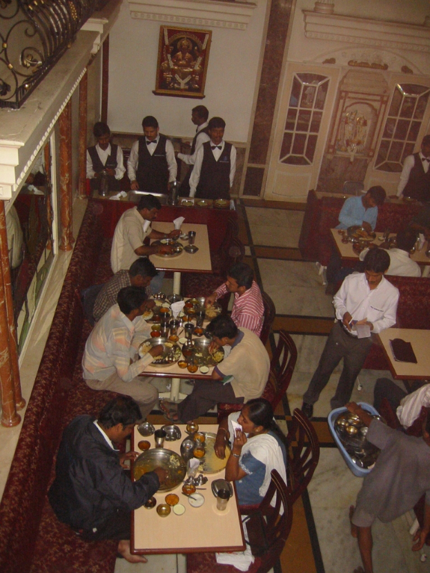 Eine Gruppe von Menschen, die an Tischen essen, mit einigen, die zum Servieren stehen, ein Foto Rahmen mit Blumen an der Wand und eine Tür im Hintergrund.
