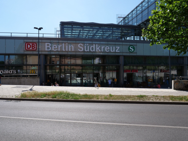 Großes Glaswand-Berliner Südkreuz-Bahnhof mit sichtbarem Text, umgeben von Straßeninfrastruktur, Fahrzeugen, Fußgängern, Fahrrädern, Bäumen und einem klaren Himmel.