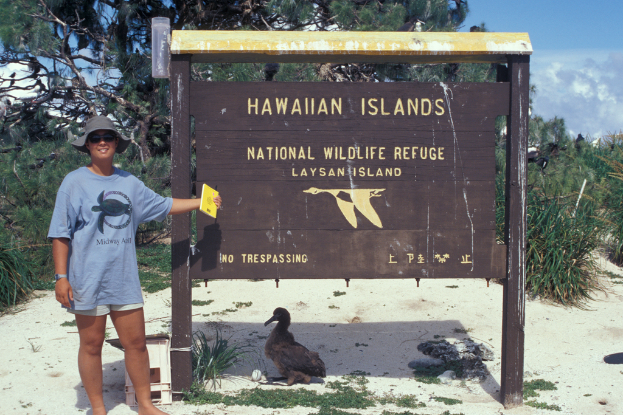 Eine Frau in einem Hut steht neben einem Plakat und hält ein Buch in der Hand, mit einer Ente auf dem Boden und Bäumen im Hintergrund unter einem klaren Himmel.