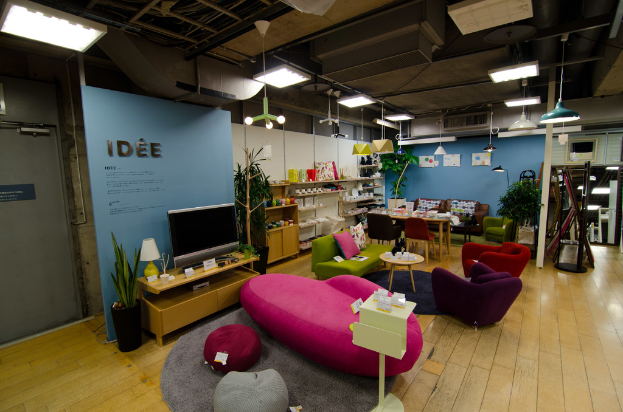 Eine Ladeneinrichtung mit Haushaltsgeräten und Möbeln, darunter ein Fernseher auf einem Tisch, eine rosafarbene Couch, ein Sofa, zwei Stühle um einen kleinen Hocker, ein Esstisch mit Stühlen, ein großes Sofa im Hintergrund und verschiedene Gegenstände auf Regalen.