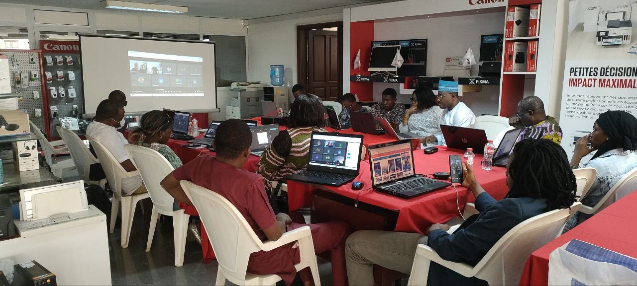 Eine Gruppe von Menschen sitzt um einen Tisch mit Laptops und anderen Gegenständen während eines digitalen Marketing-Workshops, wobei ein Banner, ein Display, eine Tür und Deckenleuchten im Hintergrund zu sehen sind.