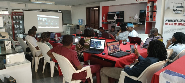 Eine Gruppe von Menschen sitzt um einen Tisch mit Laptops und anderen Gegenständen während eines digitalen Marketing-Workshops, wobei ein Banner, ein Display, eine Tür und Deckenleuchten im Hintergrund zu sehen sind.