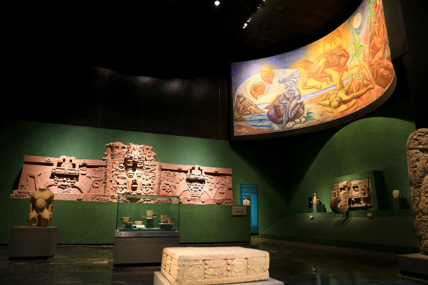 Ein Museumsraum mit verschiedenen Artefakten, darunter Skulpturen, Tischgegenstände, eine Texttafel, eine Wandmalerei und Deckenbeleuchtung im Anthropologischen Museum von Mexiko-Stadt.