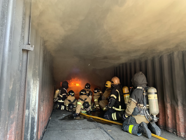 Feuerwehrleute in Helmen und Sauerstoffgeräten arbeiten in einem raucherfüllten Tunnel, einer hält ein Rohr, mit sichtbarem Feuer und einer Wand links.
