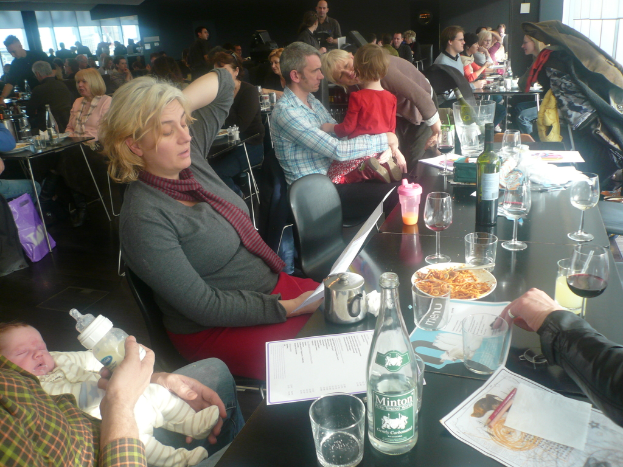 Ein Innenblick in ein belebtes Restaurant mit Menschen an Tischen, die mit Gläsern, Tellern mit Essen, Getränkeflaschen, Menükarten und anderen Gegenständen gedeckt sind.