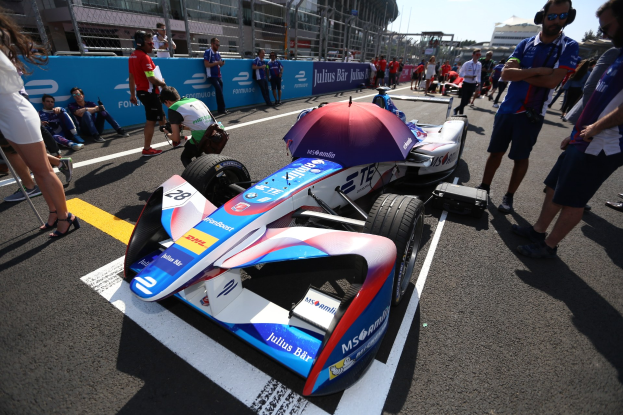 Ein Formula-E-Auto mit weißer, blauer, roter und schwarzer Lackierung steht auf einer Rennstrecke umgeben von Menschen, mit einem Schirm im Vordergrund und Gebäuden, Bäumen, Absperrungen und einem klaren blauen Himmel im Hintergrund.