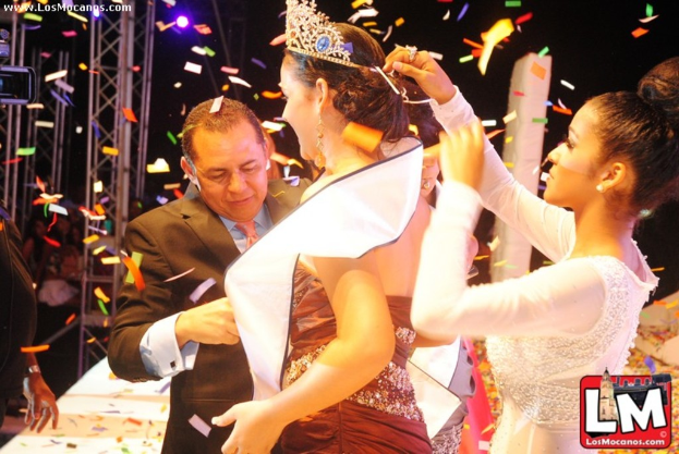 Ein Mädchen auf der Bühne bei einer Miss-World-Show mit einer Krone, dahinter ein Mädchen, das die Krone anpasst, und vorne ein Mann in einem schwarzen Anzug, der die Hand schüttelt, vor einem dunklen Hintergrund mit Metallrahmen.