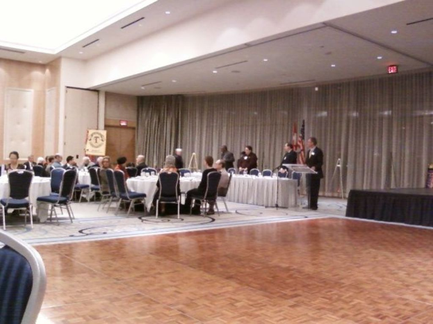 Eine Gruppe von Menschen, die auf Stühlen sitzen und stehen, mit Flaggen, Lampen, Schildern und Vorhängen im Hintergrund.