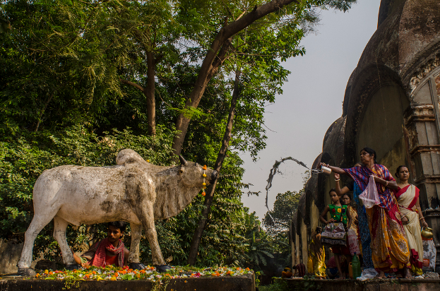 Eine Kuhskulptur mit einem Mann dahinter, umgeben von Blumen, mit ein paar Menschen daneben einem Tempel und Bäumen im Hintergrund.