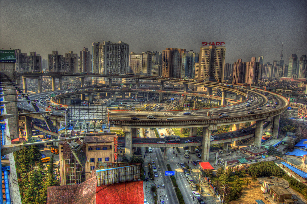Eine Stadtansicht mit einer zentralen Ringstraße mit Fahrzeugen, einer unteren Straße mit Autos und Fahrrädern, zahlreichen Hochhäusern im Hintergrund, einem Himmel oben, Bäumen links unten und kleinen Häusern, die über die Stadt verteilt sind.