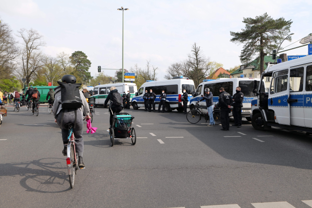 Eine Gruppe von Menschen, die auf Fahrrädern eine Straße entlangfahren, mit Polizeiwagen und anderen Fahrzeugen, einige tragen Helme und Taschen, andere schieben Kinderwagen, mit Bäumen, Laternen, Verkehrszeichen, Schildern, Gebäuden und einem bewölkten Himmel im Hintergrund.