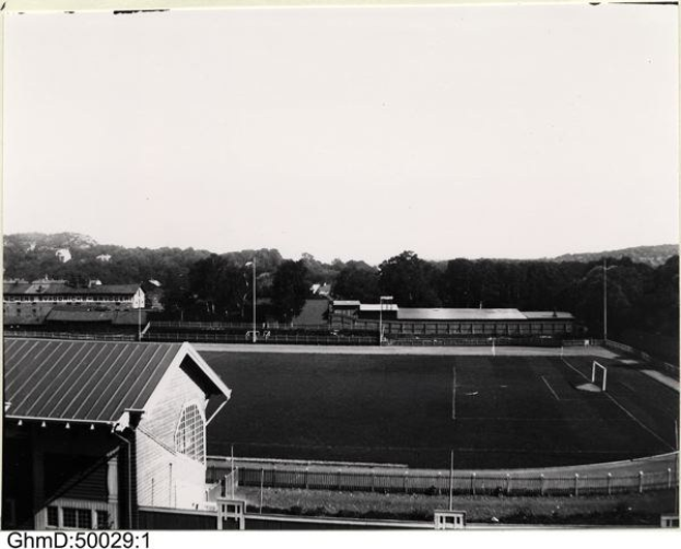 Schwarzes und weißes Foto eines Fußballfeldes mit einer Hütte auf der linken Seite, einem umgebenden Zaun, zentralen Pfosten und Bäumen mit Himmel im Hintergrund, beschriftet mit "Gillingham Football Club, 1960er Jahre" unten.