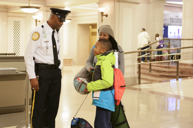 Ein Polizeibeamter in weißem Hemd, schwarzer Hose und schwarzer Mütze steht neben einem jungen Jungen, der eine Tasche hält, in einem Flughafen mit Menschen, einem Geländer, Lampen und Treppen im Hintergrund.