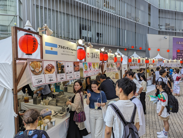Eine Gruppe von Menschen sitzt an einem Tisch mit Essen, einige tragen Taschen und halten Gläser, mit Schildern, Lichtern und Laternen im Hintergrund, was auf ein japanisches Essenfest hindeutet.