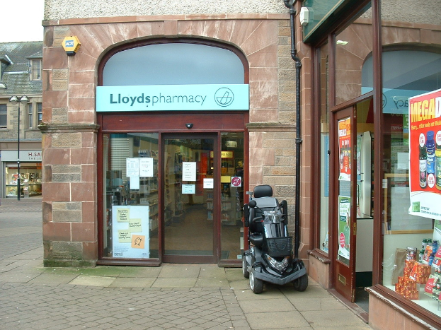 Eine Apotheke mit einem vor dem Eingang geparkten Fahrzeug und einem Gebäude in der linken Ecke.