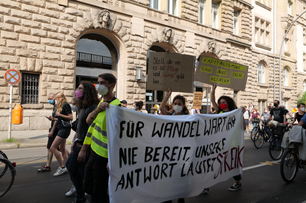 Eine Gruppe von Menschen marschiert auf einer Straße in Berlin, hält Schilder und Banner hoch und fährt mit Fahrrädern, während sie gegen die Friedensbewegung protestieren. Im Hintergrund ist ein Gebäude mit Fenstern, Bögen, Säulen und Skulpturen sowie Bäumen und einem Laternenmast zu sehen.