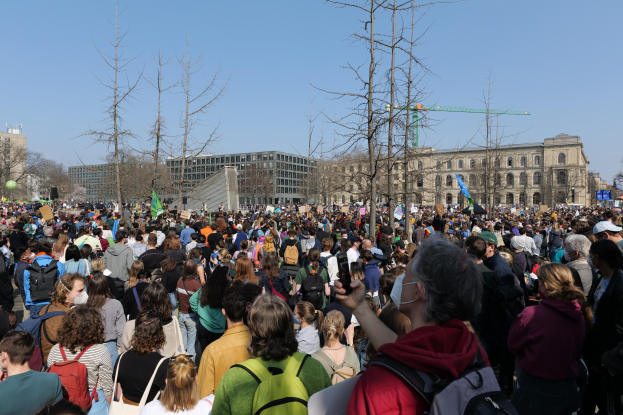 Eine große Menschenmenge nimmt an einer Klimaprotestaktion vor einem Gebäude mit Bäumen und einem klaren Himmel teil.