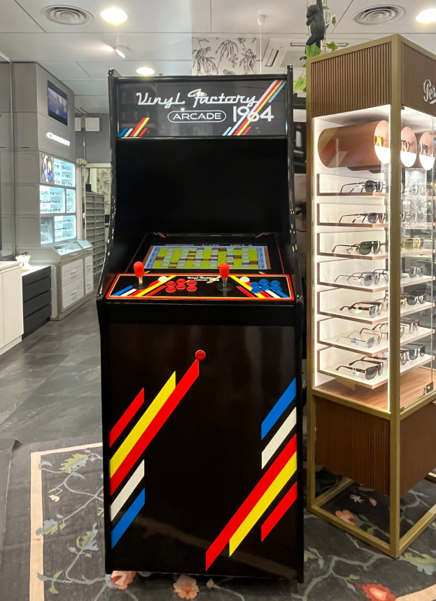 Ein Geschäft mit einem alten Arcade-Automaten in der Mitte, einem Glasvitrine auf der rechten Seite und retro-stilvollen Schränken, einem Fernseher und Deckenlampen im Hintergrund.