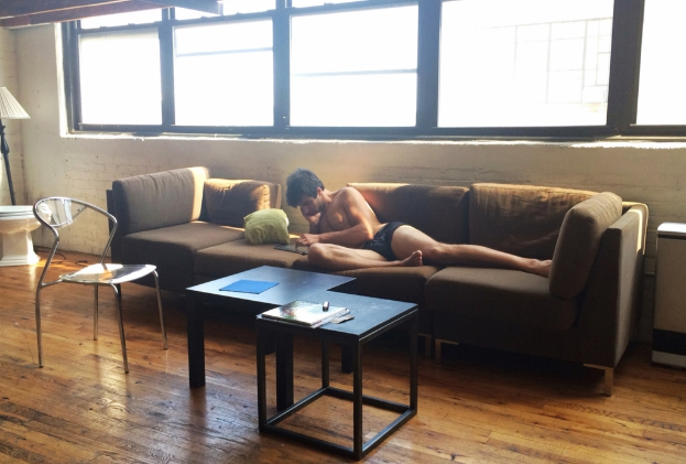 Ein Mann liegt auf einem Sofa in einem Raum mit einem Stuhl, einem Buch auf einem Tisch, einer Lampe und einem Fenster.