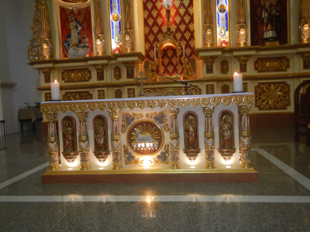 Ein Altar in einer Kirche mit Kerzen, Skulpturen, einem Mikrofon, einem Stuhl, einem Tisch mit verschiedenen Gegenständen darauf und Buntglasfenstern im Hintergrund, alles von Kerzenlicht beleuchtet.