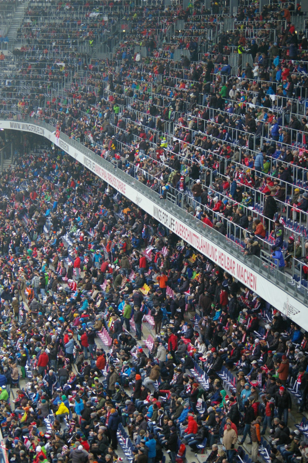Eine große Menschenmenge sitzt auf den Tribünen eines Stadions mit Geländern und Texttafeln im Hintergrund.