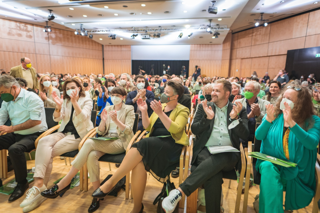 Eine Gruppe von Menschen in Stühlen klatschend, einige tragen Masken, mit Taschen auf dem Boden, vor einem Bildschirm und Deckenlichtern auf einer Coronavirus-Konferenz.