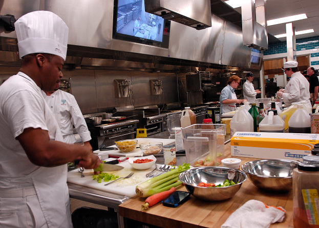Eine Gruppe von Köchen in einer Küche, mit einem im Vordergrund in einer weißen Schürze und Mütze, der ein Messer hält und von Utensilien, Schüsseln, Gemüse, einem Handy, Flaschen und anderen Gegenständen umgeben ist, während im Hintergrund ein Fernseher und Deckenleuchten zu sehen sind.