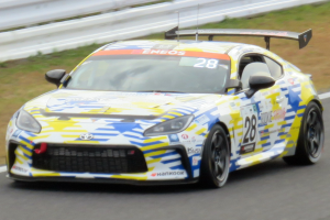 Ein Toyota Supra GT3-Rennwagen fährt auf einer Rennstrecke, mit Text und Zahlen auf dem Auto, einer Begrenzung im Hintergrund und Gras am Boden.