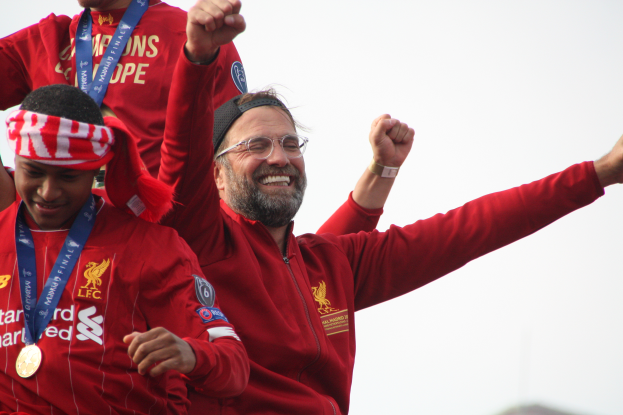 Liverpool-Trainer Jürgen Klopp feiert mit seinen Spielern den UEFA-Champions-League-Sieg, während er eine rote Jacke trägt und von zwei medaillentragenden Personen umgeben ist. Der Himmel ist im Hintergrund zu sehen.